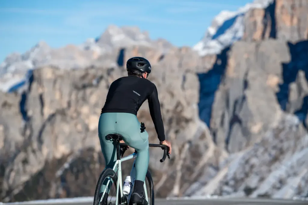 Un ciclista, que lleva un maillot negro de manga larga y casco, pedalea por una carretera de montaña con picos rocosos al fondo.
