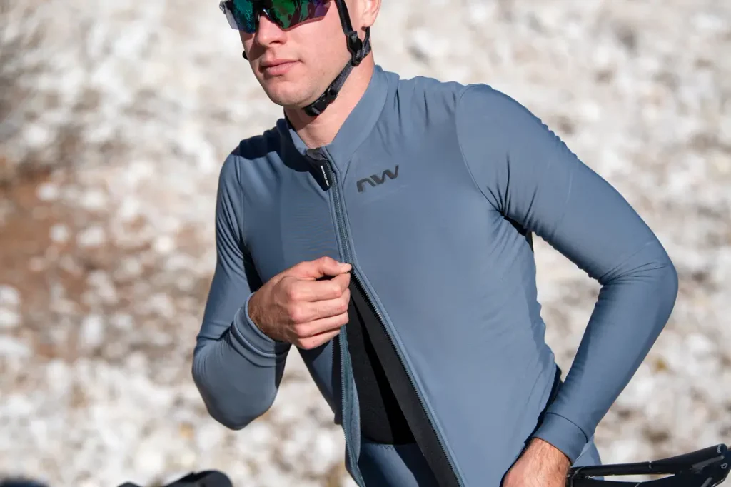 Una persona que lleva gafas de sol y casco se abrocha la cremallera de una chaqueta de ciclista azul con las letras «NW», de pie al aire libre.