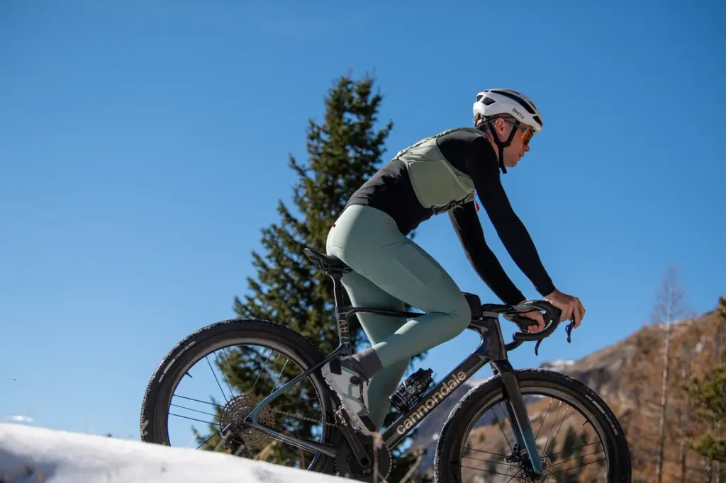 Un ciclista, que lleva casco y ropa de ciclismo, circula en una bicicleta Cannondale en un día despejado, con un cielo azul y árboles al fondo.