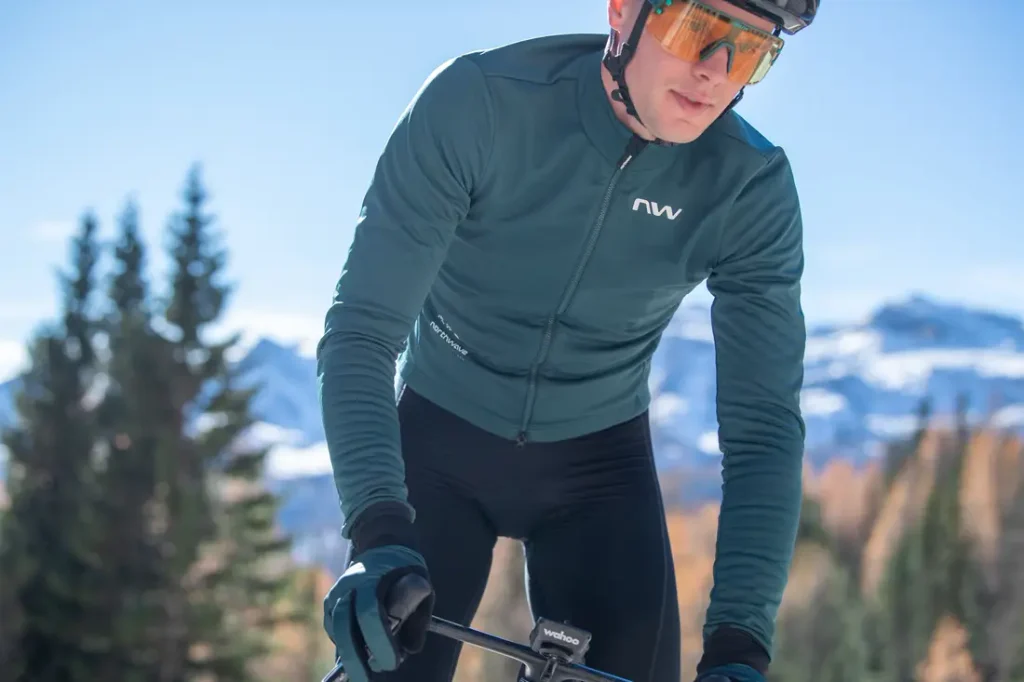 Un ciclista que lleva gafas de sol, guantes y una chaqueta de color verde azulado va en bicicleta al aire libre, con montañas y árboles al fondo.