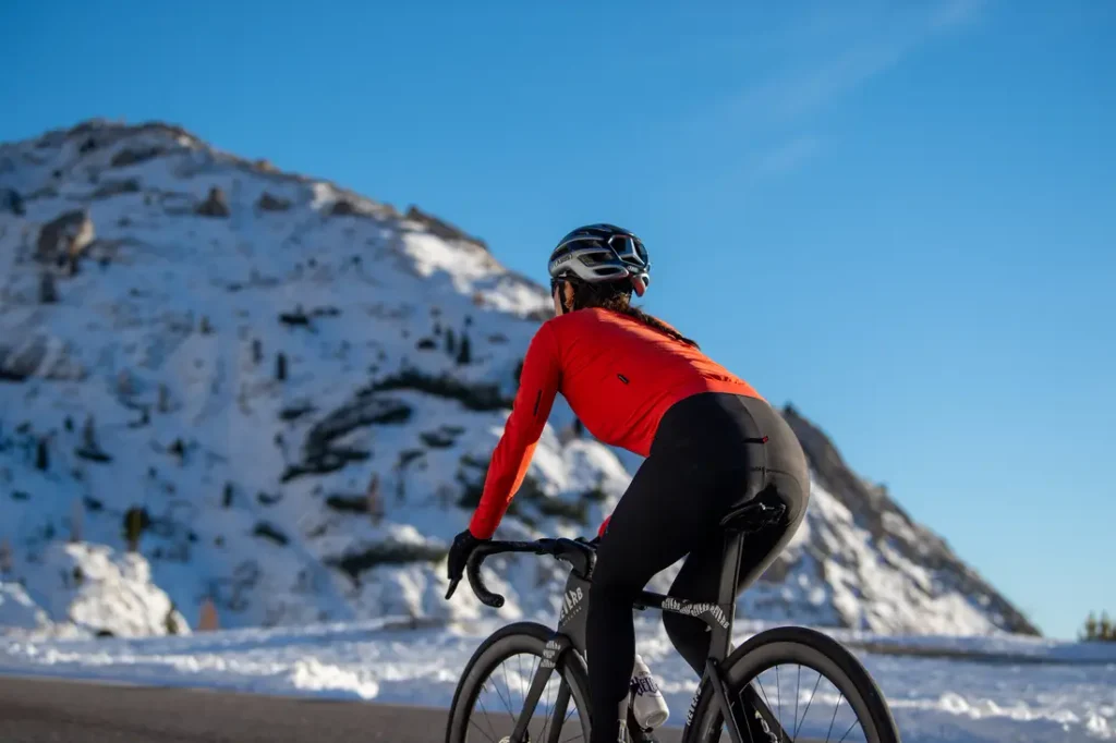 Un ciclista, con una chaqueta roja y casco, circula en bicicleta de carretera por una carretera asfaltada, con una montaña cubierta de nieve al fondo.