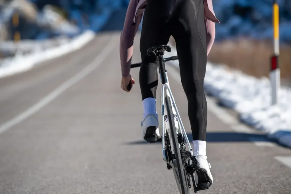 Un ciclista, que lleva mallas negras y zapatillas blancas, va en bicicleta de carretera por una carretera asfaltada con nieve a los lados.