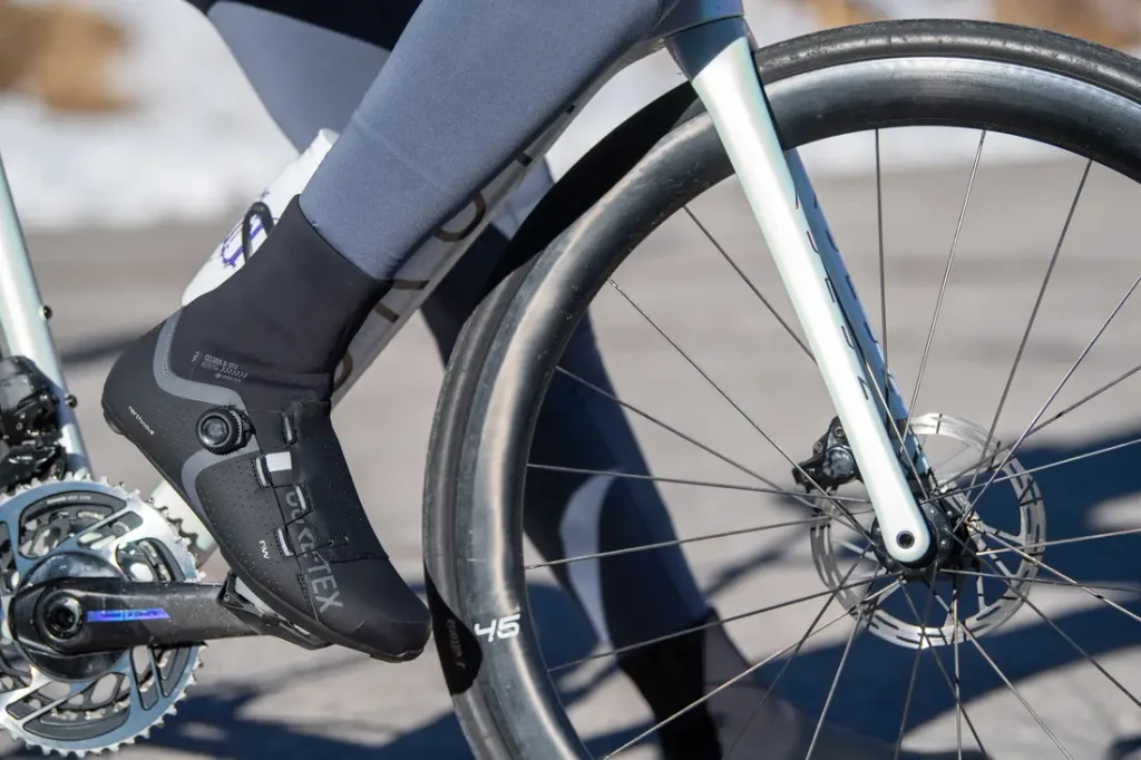 Primer plano de una persona montando en bicicleta, en el que se ve la zapatilla negra de ciclismo del ciclista enganchada al pedal y parte de la rueda delantera y la horquilla de la bicicleta.