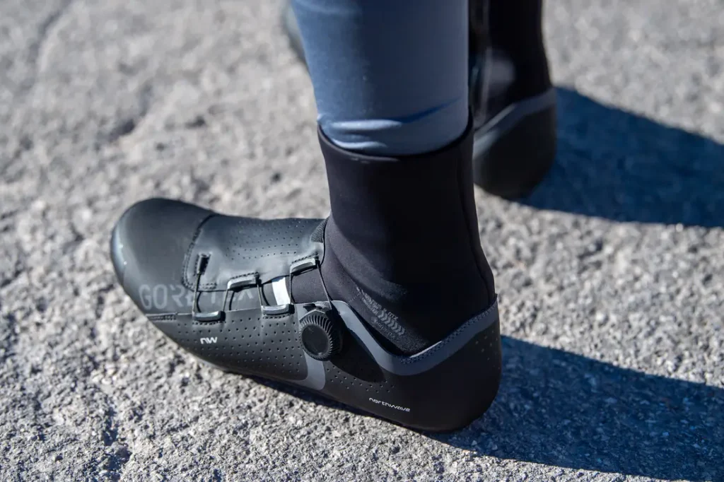Una persona que lleva unas zapatillas de ciclismo negras con cierre de dial y unos leggings de color gris azulado está de pie sobre una superficie de asfalto irregular.