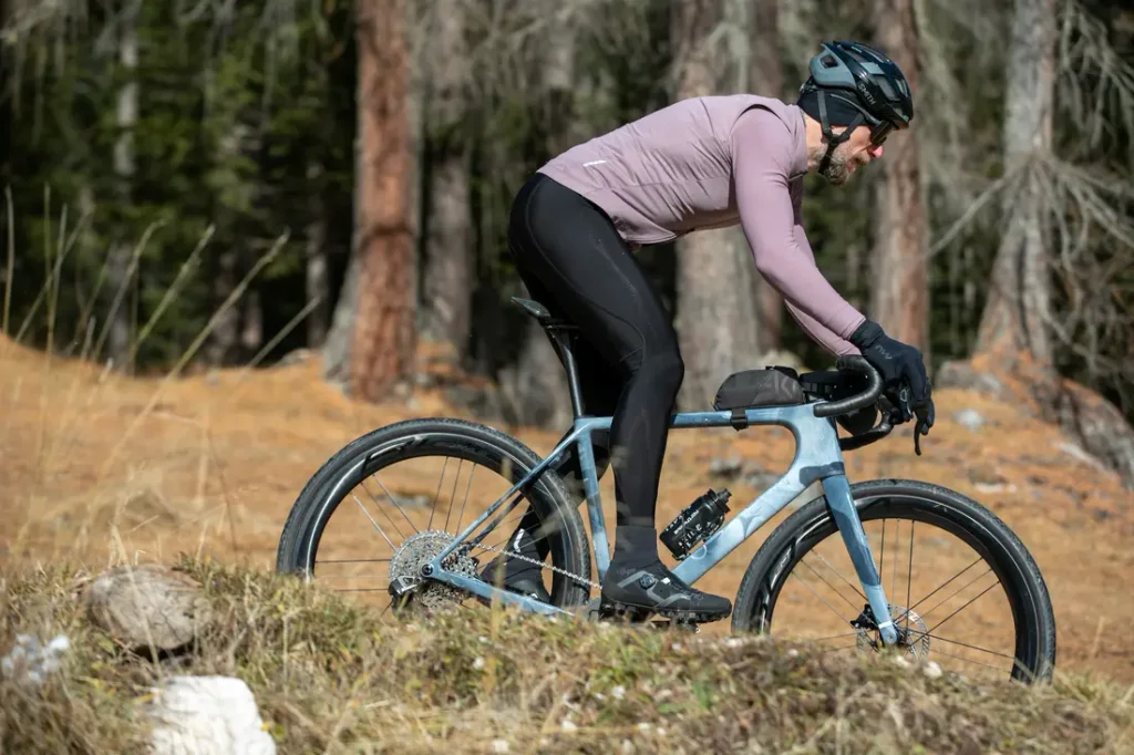 Una persona que lleva casco, guantes y ropa de ciclismo pedalea con una bicicleta de gravel cuesta arriba por un sendero cubierto de hierba, con árboles al fondo.