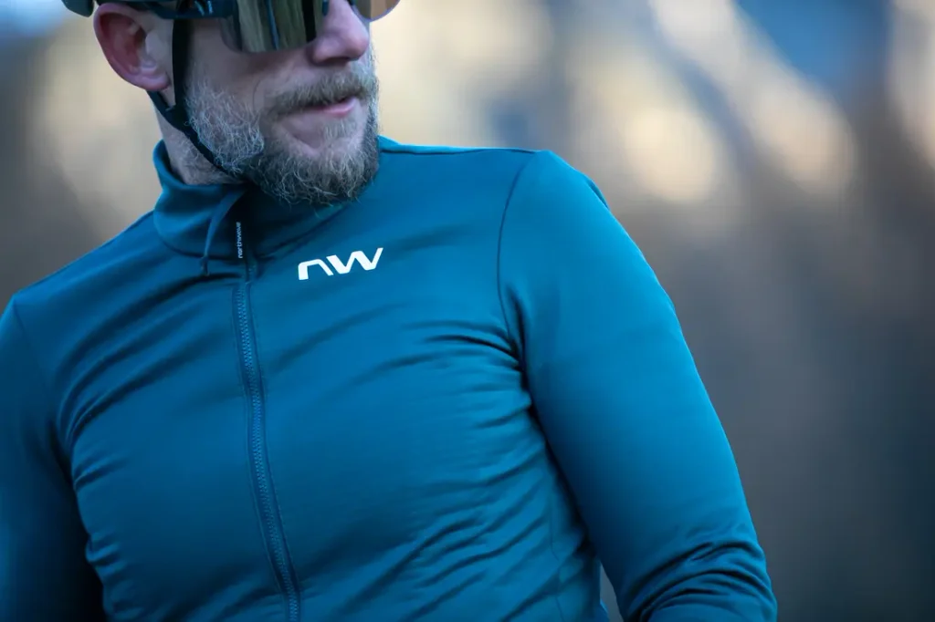 Un hombre que lleva gafas de sol y un maillot de ciclismo azul de manga larga con el logotipo «nw» en blanco está de pie al aire libre.