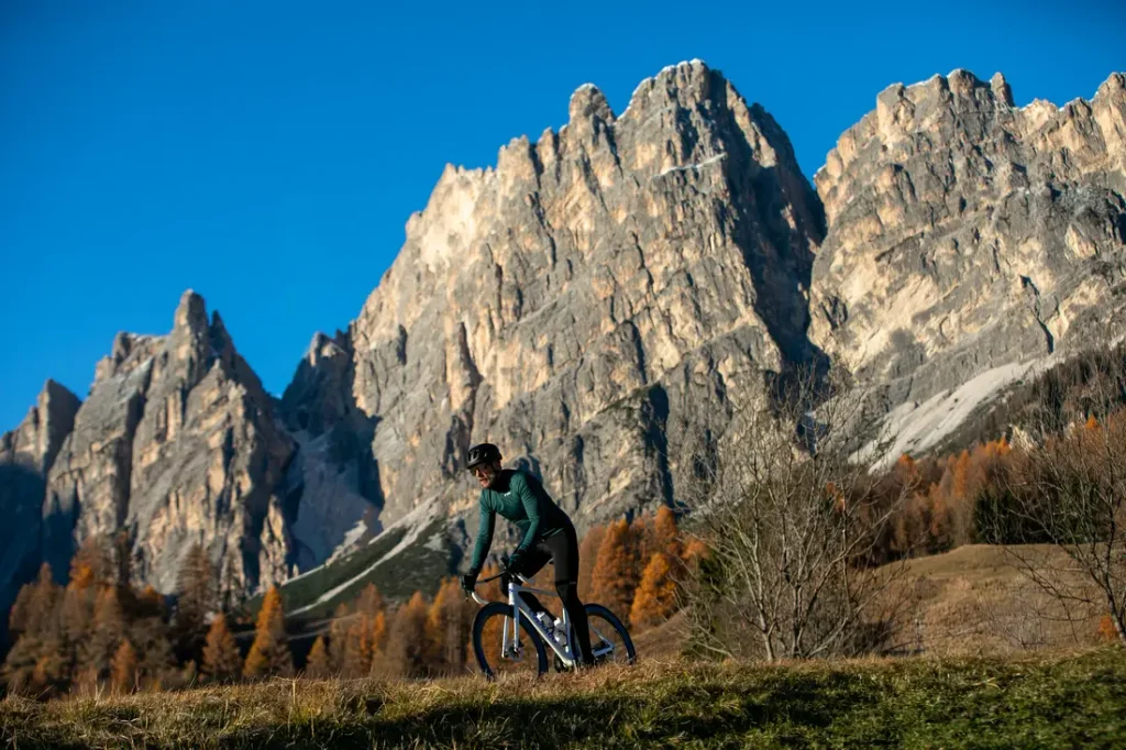 Una persona va en bicicleta por un camino cubierto de hierba, con altas montañas rocosas y un cielo azul y despejado al fondo.