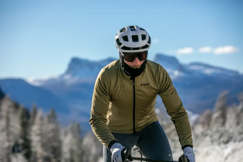 Un ciclista, que lleva casco, gafas de sol y chaqueta, pedalea al aire libre con montañas y árboles al fondo.