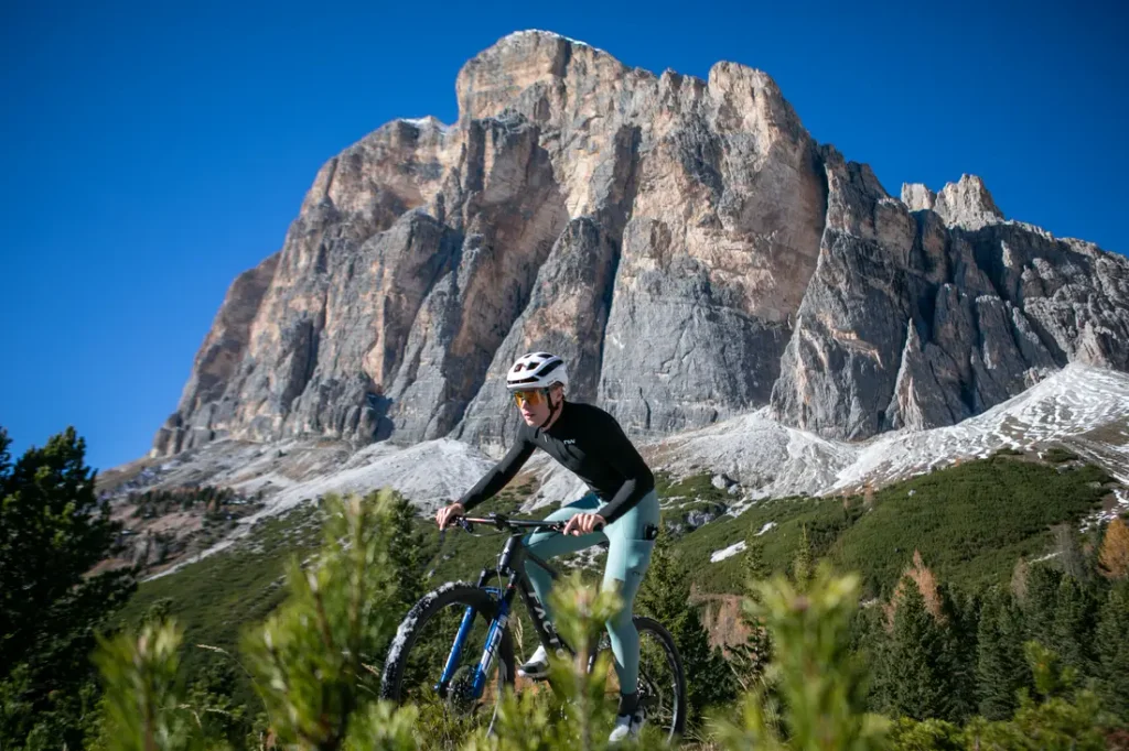 Un ciclista, con casco y gafas de sol, va en bicicleta de montaña por un sendero, con una gran montaña rocosa y un cielo azul y despejado al fondo.