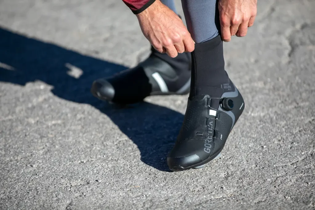 Una persona se coloca unos cubrezapatillas negros de ciclismo sobre las zapatillas mientras está de pie sobre una superficie pavimentada.