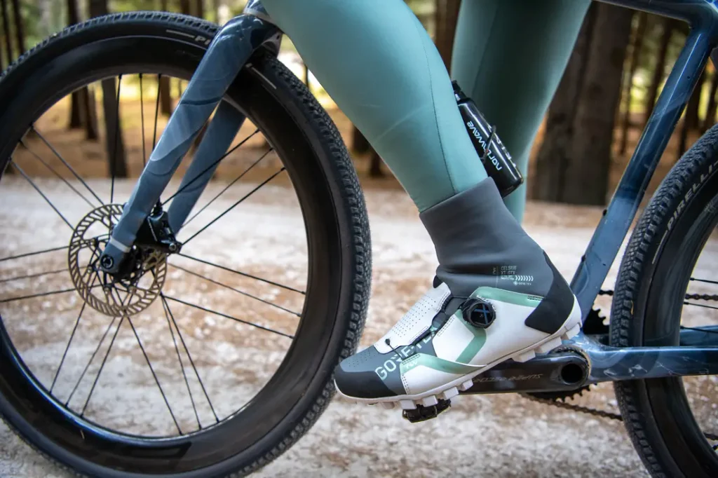 Primer plano de una persona que lleva mallas verdes y zapatillas de ciclismo, pedaleando en una bicicleta por un camino de tierra que atraviesa una zona boscosa.