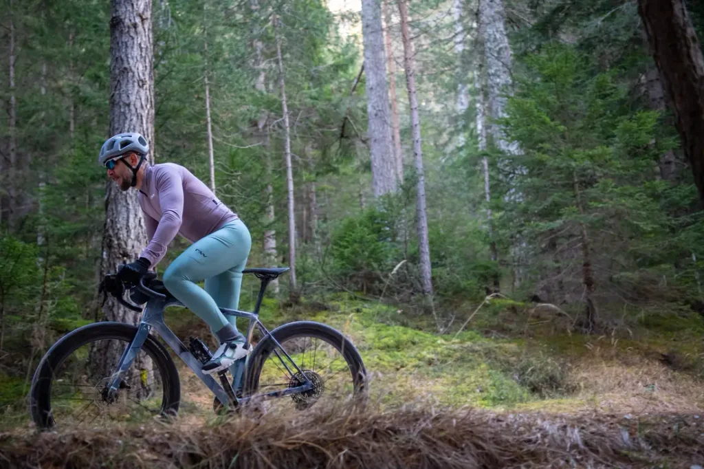 Una persona que lleva casco y gafas de sol va en bicicleta por un sendero de tierra que atraviesa un bosque con árboles altos y frondosa vegetación.