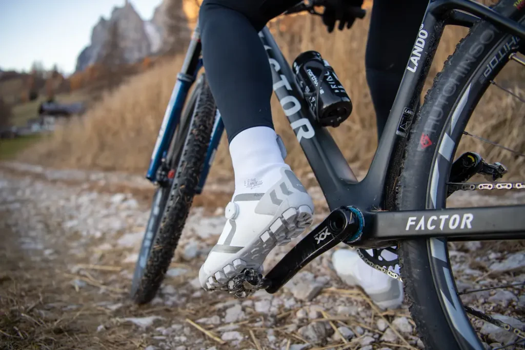 Primer plano del pie de un ciclista, calzado con una zapatilla blanca de ciclismo, sobre el pedal de una bicicleta Factor negra, mientras circula por un sendero rocoso.