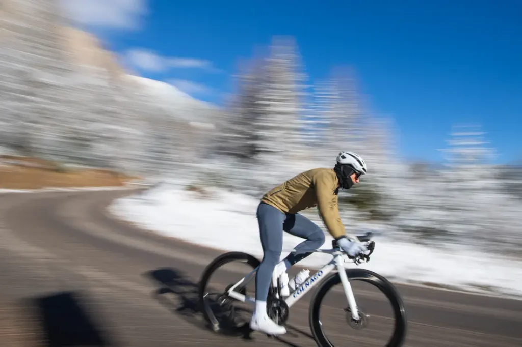 Una persona que lleva casco y ropa de invierno va en bicicleta a gran velocidad por una carretera asfaltada, con árboles cubiertos de nieve al fondo.