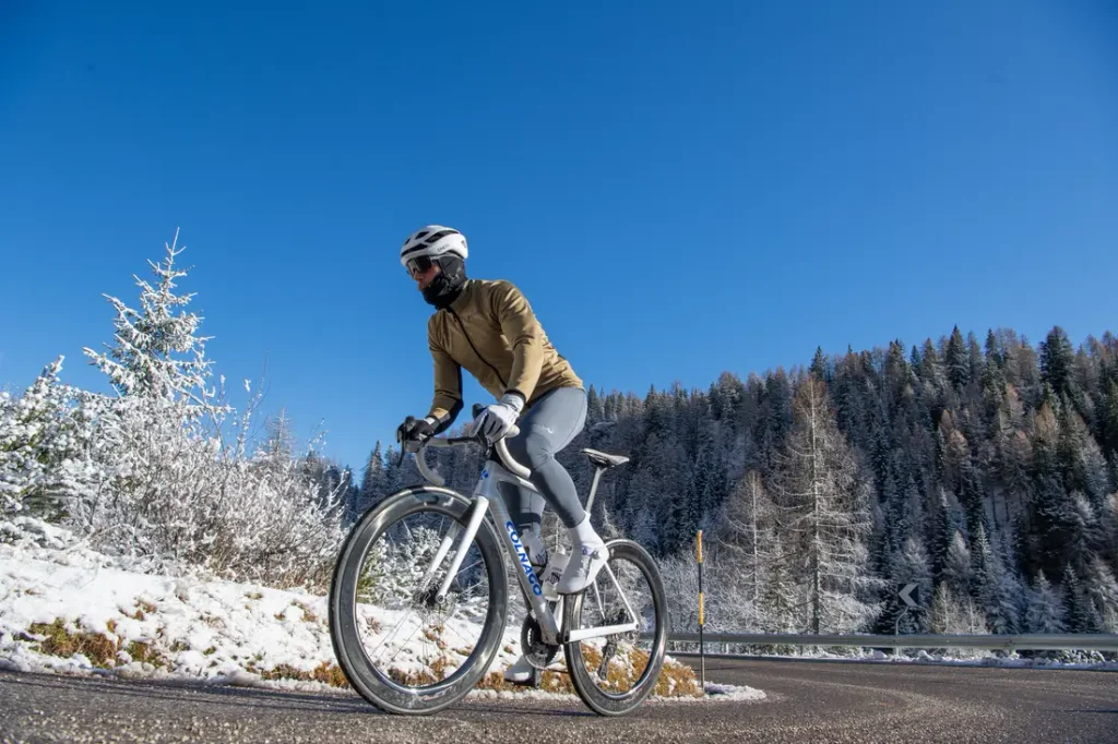 Una persona que lleva casco y ropa de ciclismo de invierno va en bicicleta de carretera por una carretera asfaltada, con árboles cubiertos de nieve y un cielo azul despejado al fondo.