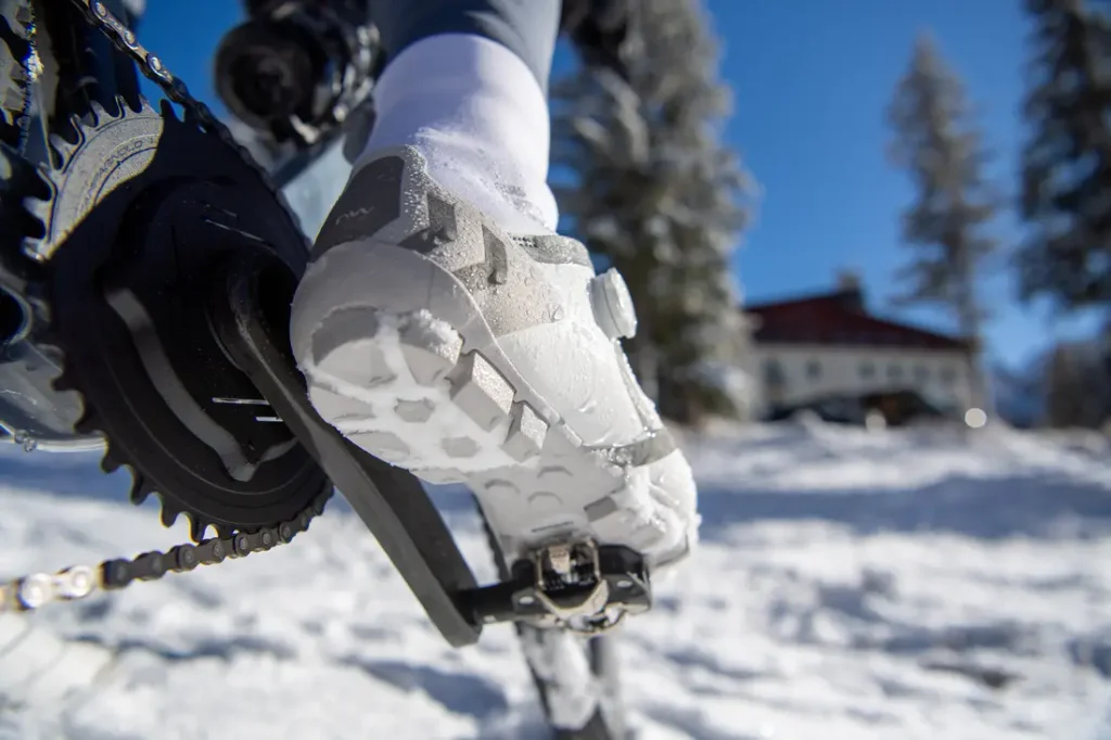 Primer plano de la zapatilla de un ciclista enganchada al pedal de una bicicleta, con el suelo cubierto de nieve y una casa y unos árboles visibles en el fondo borroso.