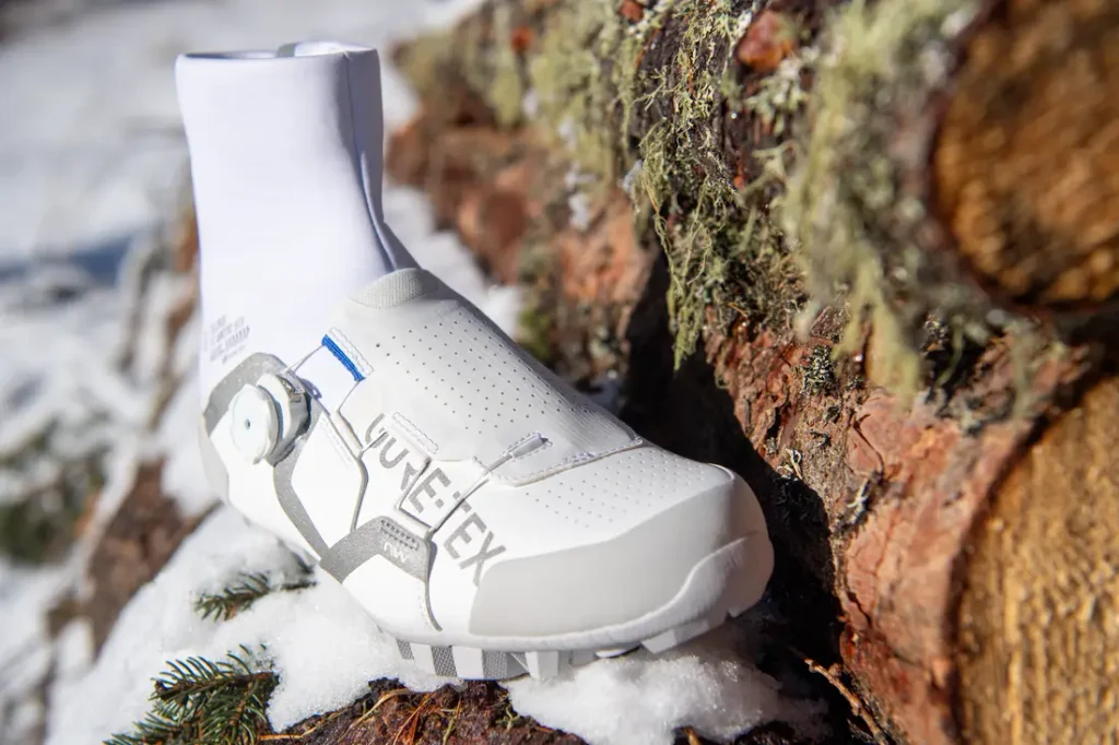 Una zapatilla de ciclismo blanca, con un calcetín alto hasta el tobillo, descansa sobre una pila de troncos cubiertos de una ligera capa de nieve; presenta la palabra «VULCLEX» en el lateral y un sistema de cierre con rueda giratoria.