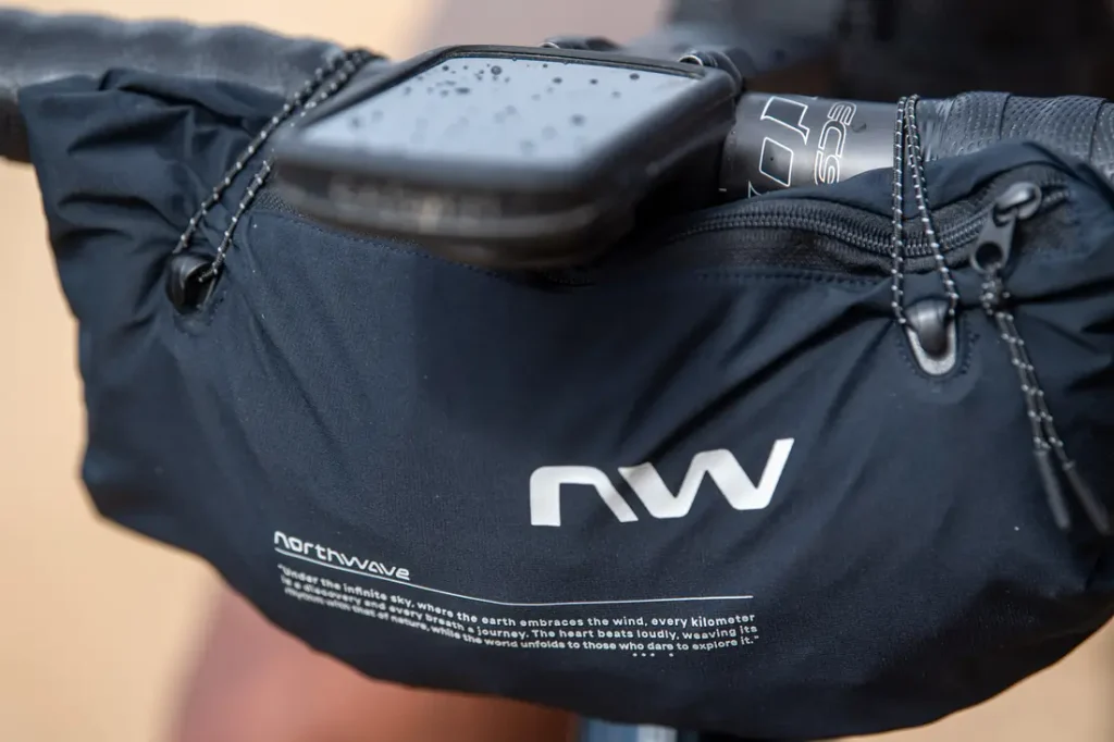 Un primer plano de una bolsa negra para manillar de bicicleta de la marca Northwave con letras blancas, fijada al manillar, con un smartphone colocado encima, en el que se ven gotas de agua sobre el teléfono.