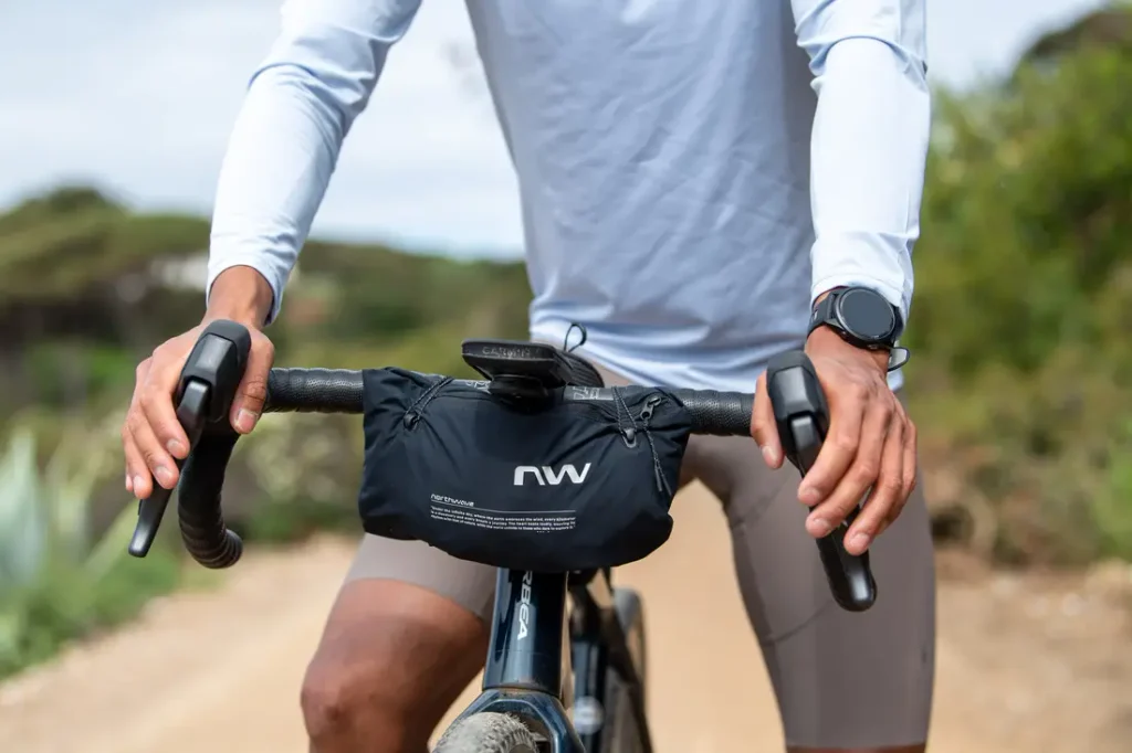 Persona vestida con una camiseta blanca de manga larga y que lleva un reloj, montada en bicicleta por un camino de tierra, con las manos agarradas al manillar, en el que hay una pequeña bolsa negra.