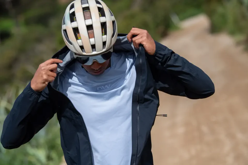 Una persona que lleva casco y gafas de sol se abrocha la cremallera de una chaqueta oscura sobre una camisa blanca mientras está de pie en un camino de tierra al aire libre.
