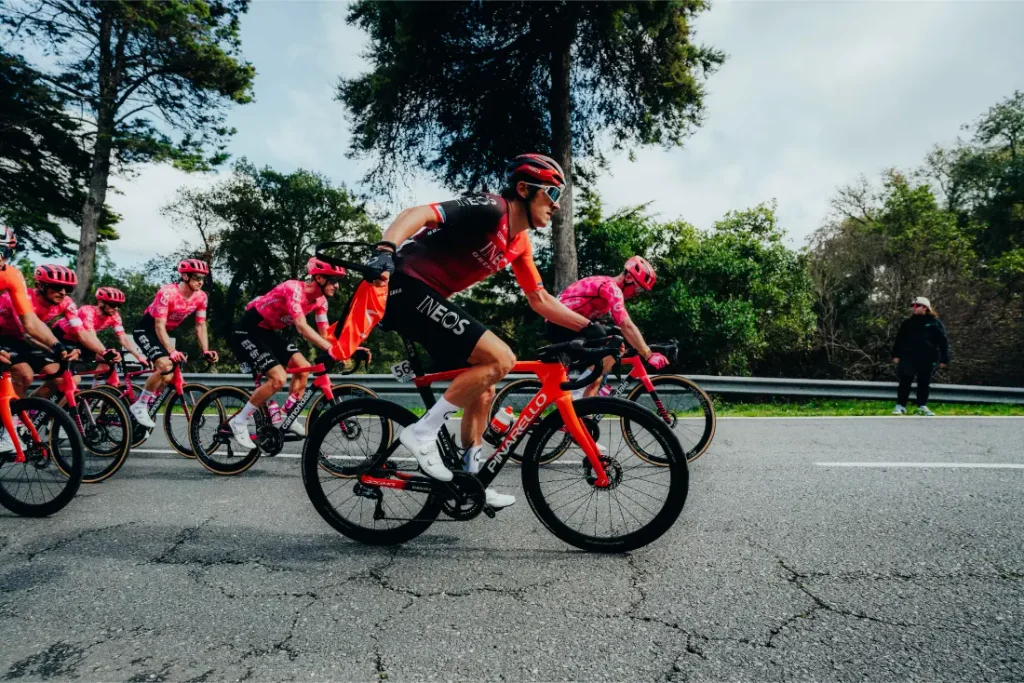 Un grupo de ciclistas, con casco y equipación del equipo, pedalea por una carretera bordeada de árboles durante una carrera; el ciclista que va en cabeza lleva la inscripción «INEOS».