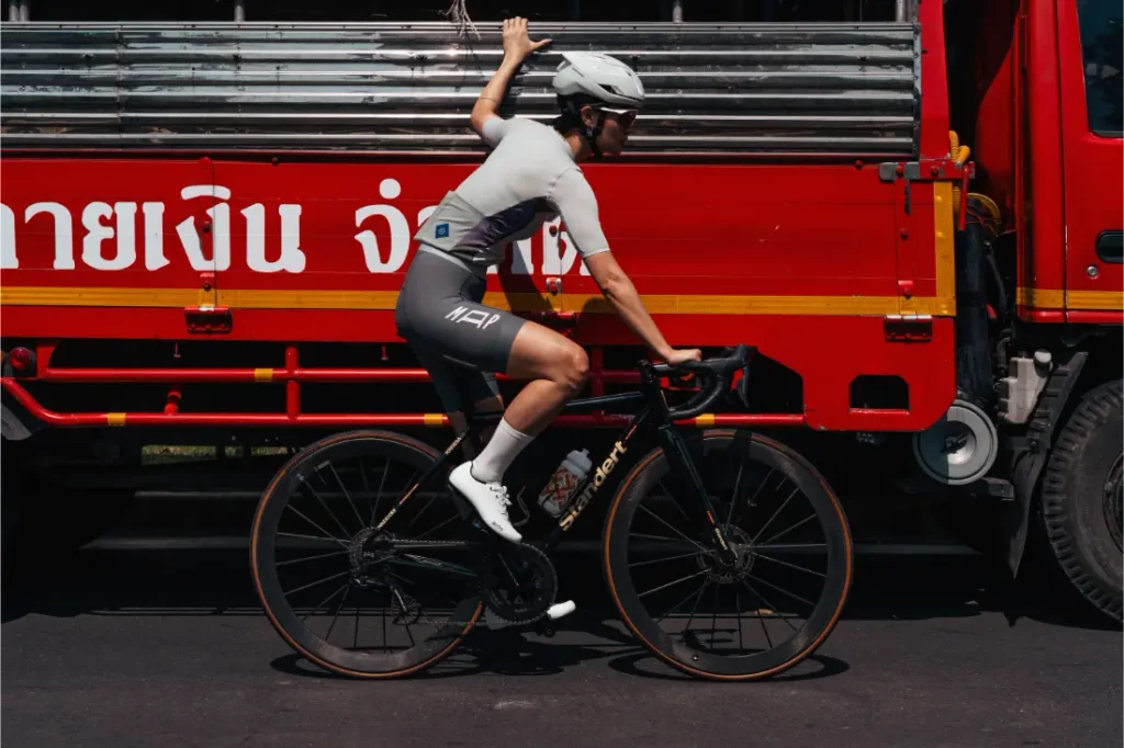 Un ciclista, con casco y ropa de competición, pasa en una bicicleta negra junto a un camión rojo, agarrándose con una mano al costado del camión.