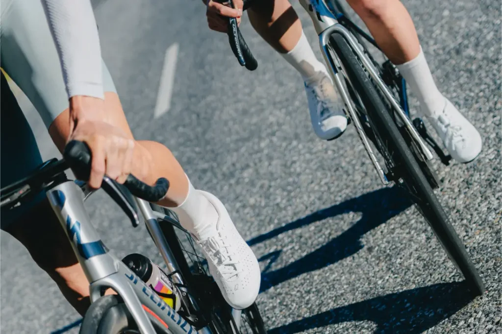 Dos ciclistas, con zapatillas y calcetines blancos, circulan en bicicleta de carretera por una calle asfaltada bañada por el sol; solo se ven la parte inferior de sus cuerpos y algunos detalles de las bicicletas.
