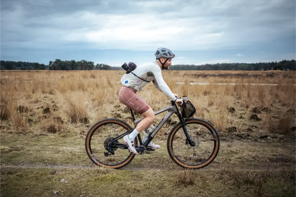 Un ciclista, con casco y ropa deportiva, va en bicicleta por un camino de tierra que atraviesa un campo abierto y cubierto de hierba, bajo un cielo nublado.