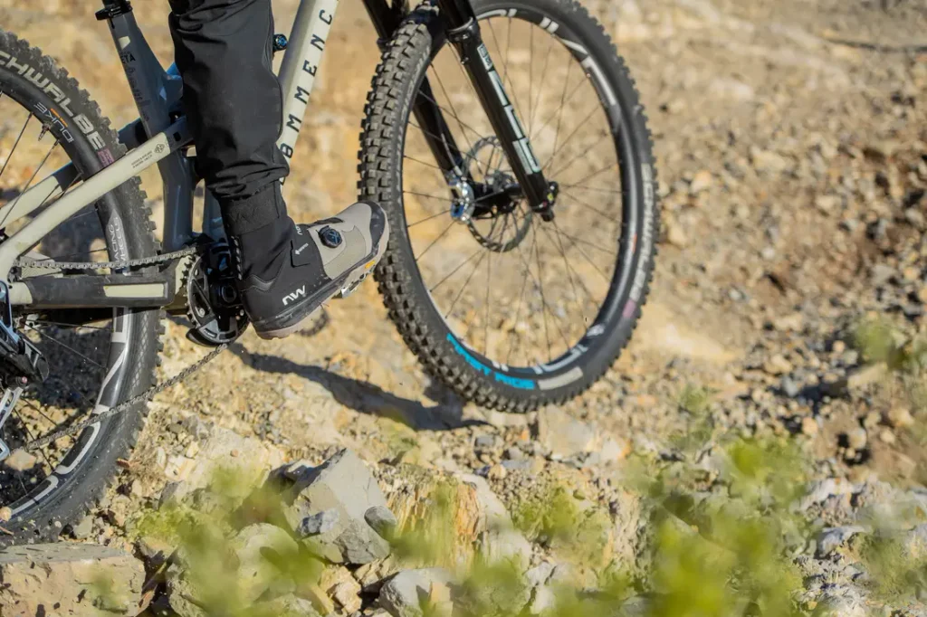 Una persona montando en bicicleta de montaña por un sendero rocoso, en la que se aprecian las ruedas y los pedales de la bicicleta, así como la parte inferior de las piernas del ciclista, que lleva ropa negra.