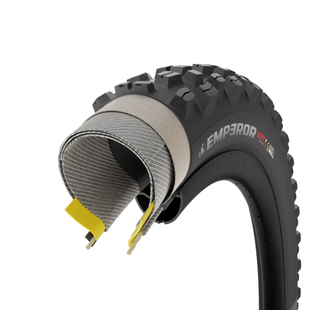 Una sección transversal de una cubierta de bicicleta negra y con protuberancias, con la inscripción «EMPEROR SOFT», muestra las capas internas y los detalles de su construcción, lo que recuerda el diseño robusto de los neumáticos Pirelli Scorpion para MTB.