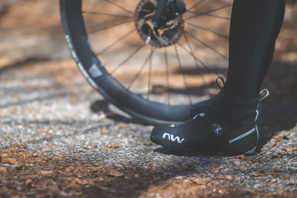 Una persona que lleva zapatillas de ciclismo negras está de pie junto a una rueda de bicicleta en un sendero iluminado por el sol y cubierto de hojas.