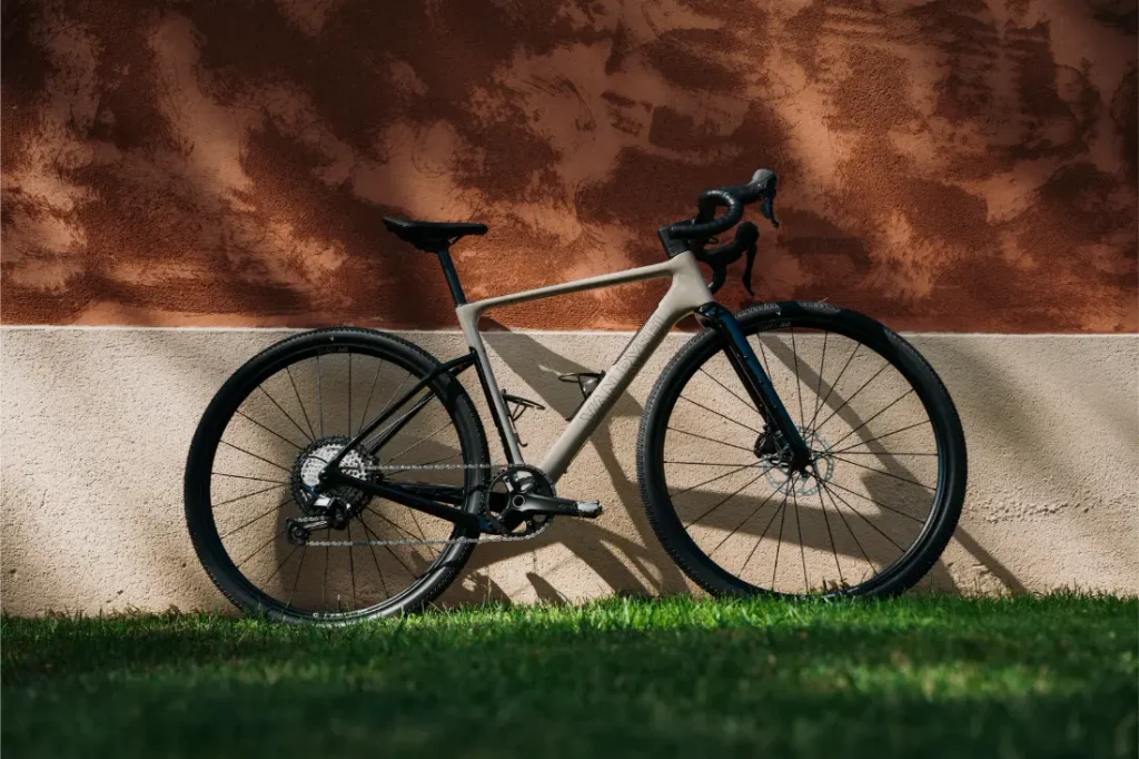 Una moderna bicicleta de carretera Van Rysel RCR-G, con manillar de carretera y neumáticos finos, se apoya contra una pared texturizada en dos tonos, proyectando una sombra sobre el césped que hay debajo; lista tanto para el asfalto liso como para explorar por caminos de grava suelta.