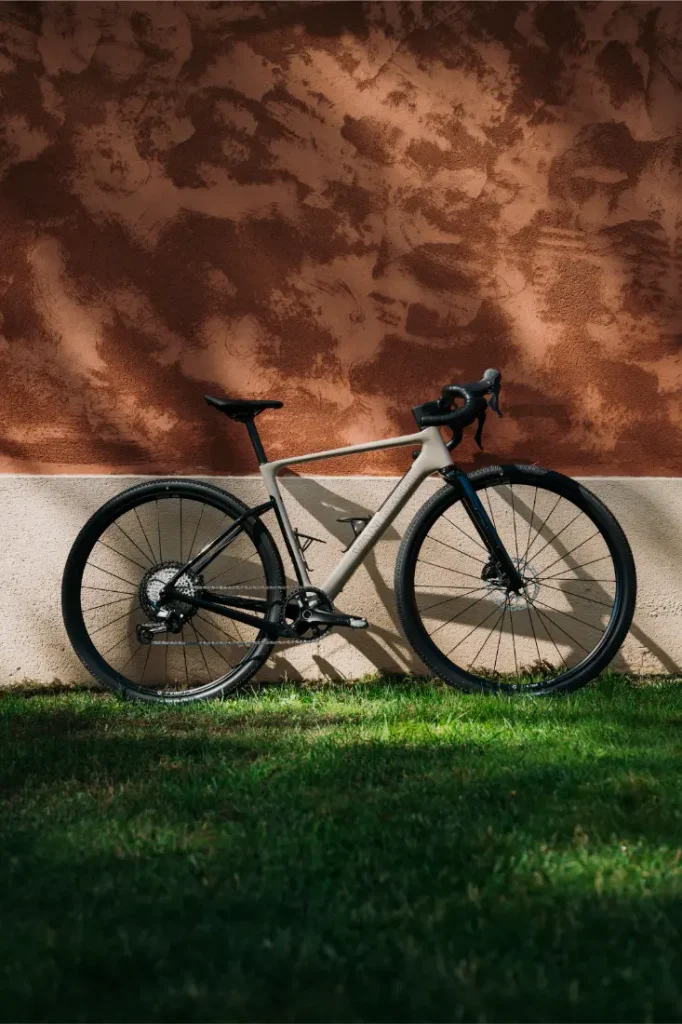 Una bicicleta de carretera gris con neumáticos negros está aparcada sobre la hierba, frente a una pared de estuco de color marrón rojizo con textura, a la luz del sol.