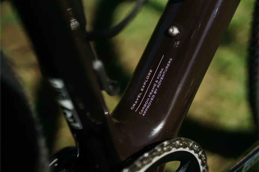 Primer plano de un cuadro de bicicleta Van Rysel RCR-G de color marrón, en el que se aprecia, cerca del juego de bielas, el texto «Gravel Explore, cuadro de carbono, diseñado por aventureros».