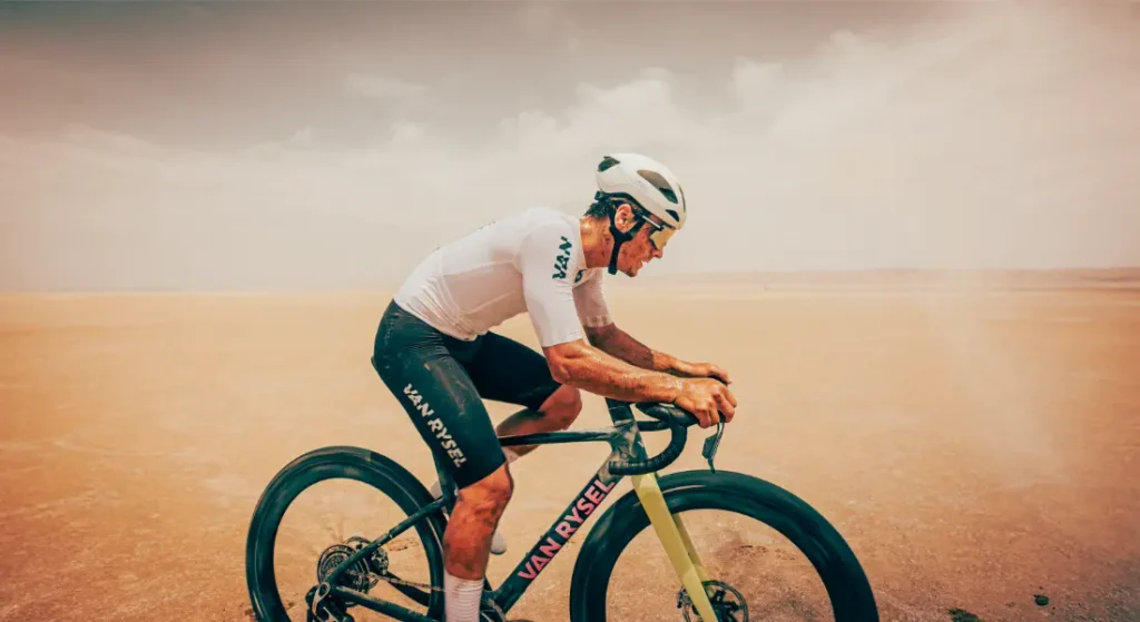 Un ciclista, con casco y maillot blancos, recorre un paisaje seco y arenoso a lomos de una bicicleta de gravel Van Rysel RCR-G, bajo un cielo brumoso.