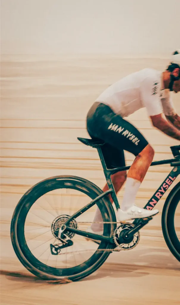 Un ciclista, vestido con ropa de competición, pedalea en una bicicleta Van Rysel RCR-G sobre una superficie de grava arenosa y difuminada, lo que resalta el movimiento y la velocidad.