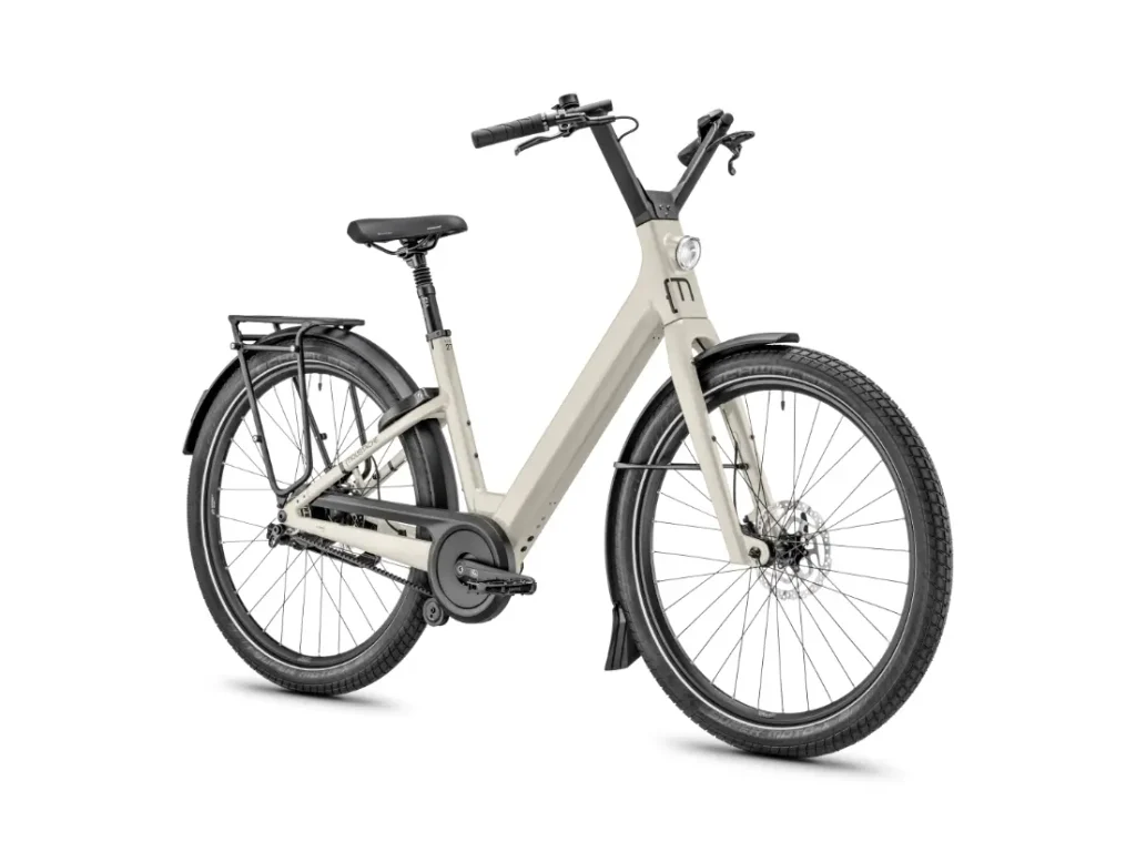 Una bicicleta eléctrica de cuadro bajo de color beige, la Moustache Lundi 27, con faro delantero, portaequipajes trasero y guardabarros negros —perfecta para explorar la ciudad—, representada sobre un fondo blanco.