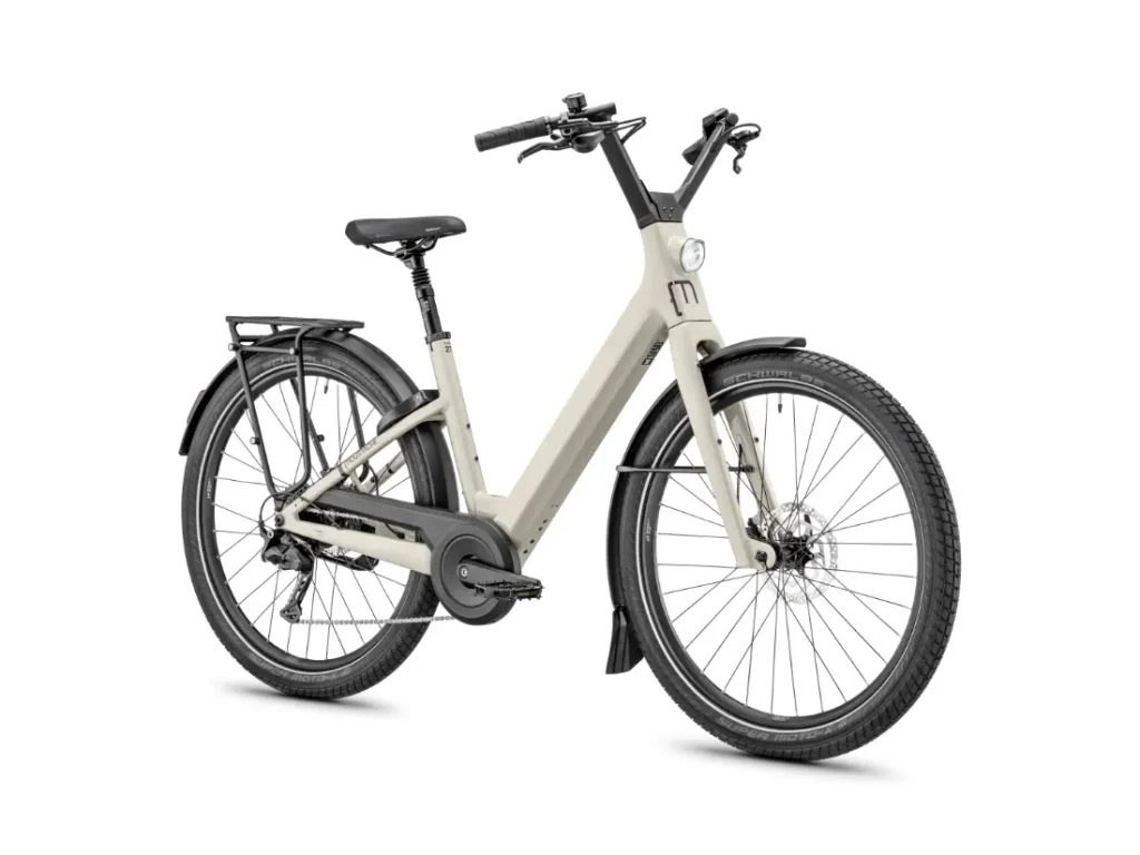 Una bicicleta eléctrica Moustache Lundi 27 de color beige, con cuadro abierto, diseñada para la ciudad, que cuenta con guardabarros delanteros y traseros, un portaequipajes trasero, luz delantera y manillar ergonómico, mostrada sobre un fondo blanco.