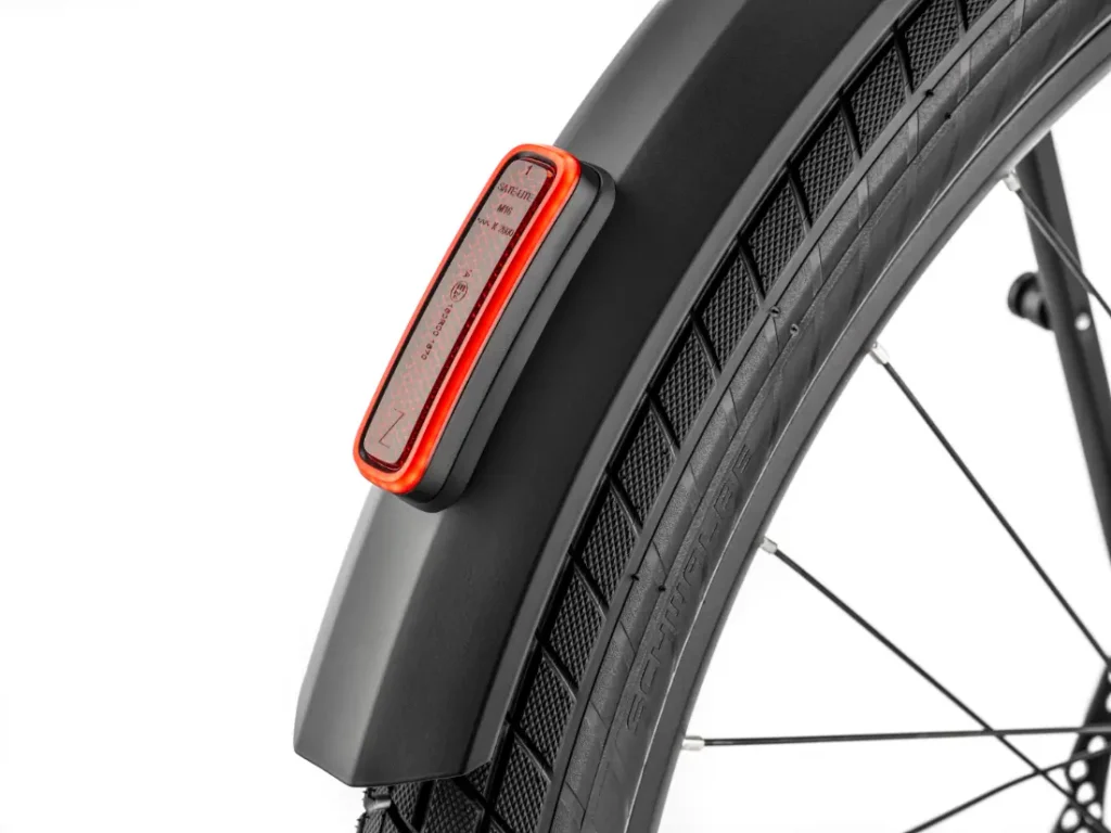 Primer plano del guardabarros trasero de una bicicleta «Moustache Lundi 27» con una luz trasera LED roja incorporada y parte de una rueda negra con la banda de rodadura y los radios visibles, ideal para moverse por la ciudad.