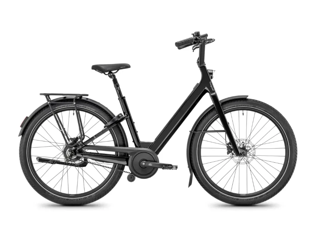 Una bicicleta urbana Moustache Lundi 27 de color negro, con cuadro abierto, portaequipajes trasero, guardabarros y frenos de disco, fotografiada de perfil sobre un fondo blanco: perfecta para desplazarse con estilo por la ciudad.