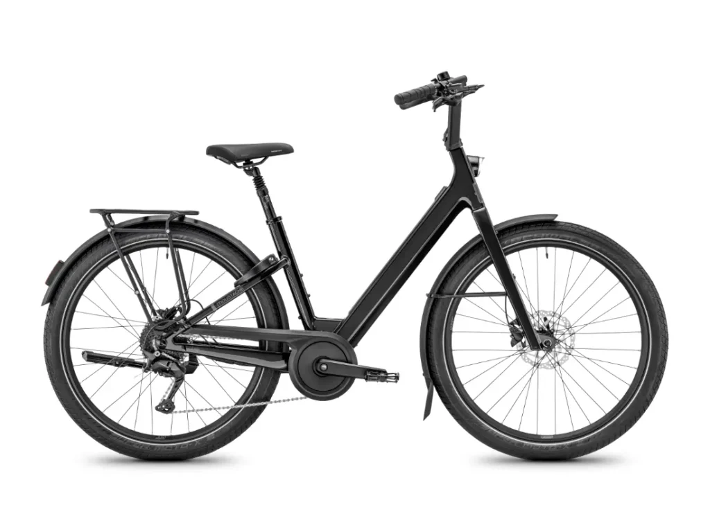 Se muestra de perfil, sobre un fondo blanco, una bicicleta eléctrica urbana negra, la Moustache Lundi 27, con cuadro de acceso fácil, portaequipajes trasero y frenos de disco: perfecta para moverse por la ciudad con comodidad y estilo.
