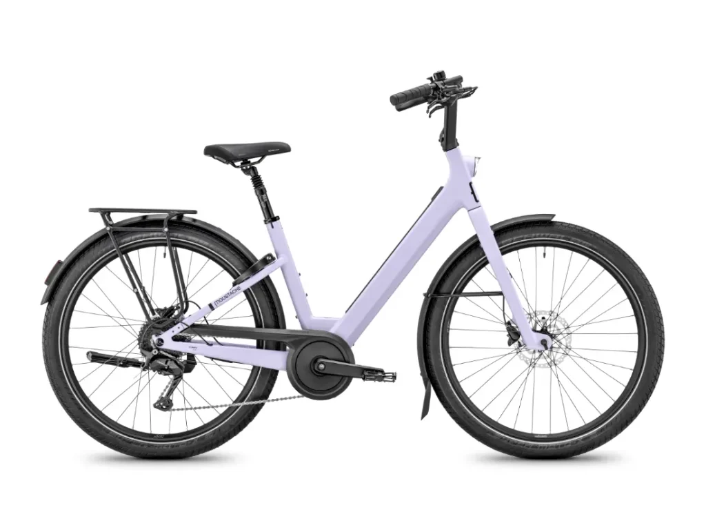 Una bicicleta urbana de cuadro abierto de color morado claro, inspirada en el diseño de la Moustache Lundi 27, con portaequipajes trasero, sillín negro y manillar recto —perfecta para moverse por la ciudad—, vista de perfil sobre un fondo blanco.