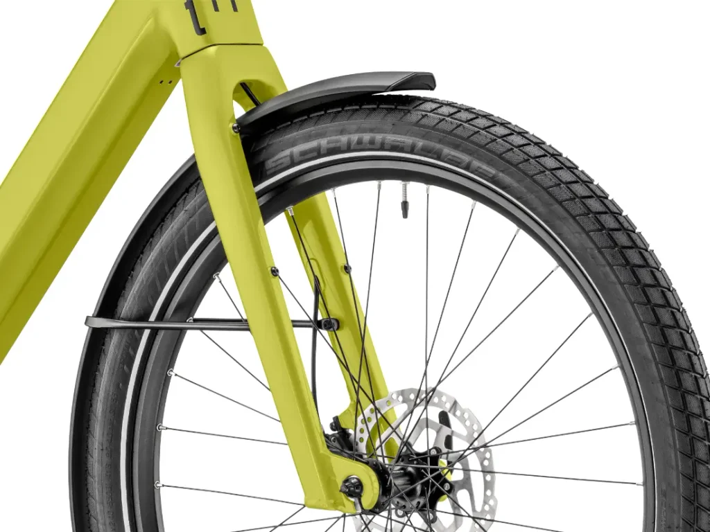 Primer plano de la rueda delantera de una bicicleta amarilla «Moustache Lundi 27», en el que se aprecian el neumático, los radios, el freno de disco, el guardabarros y parte del cuadro: perfecta para moverse por la ciudad con estilo.