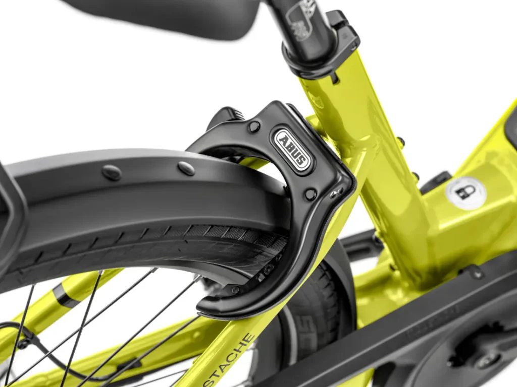 Primer plano de un candado de anillo negro de la marca ABUS fijado a la rueda trasera y al cuadro de una bicicleta amarilla Moustache Lundi 27, perfecta para moverse por la ciudad con total seguridad.