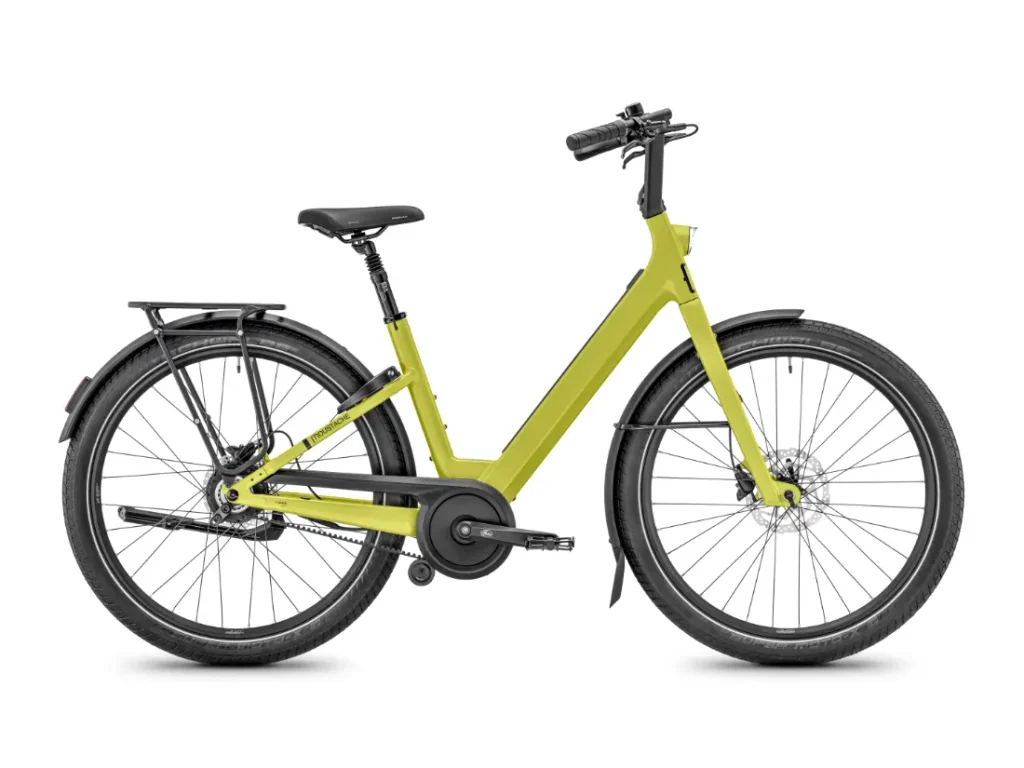 Una bicicleta eléctrica Moustache Lundi 27 de barra baja de color amarillo, diseñada para los desplazamientos urbanos, con guardabarros, portaequipajes trasero y frenos de disco delanteros y traseros, mostrada de perfil sobre un fondo blanco.