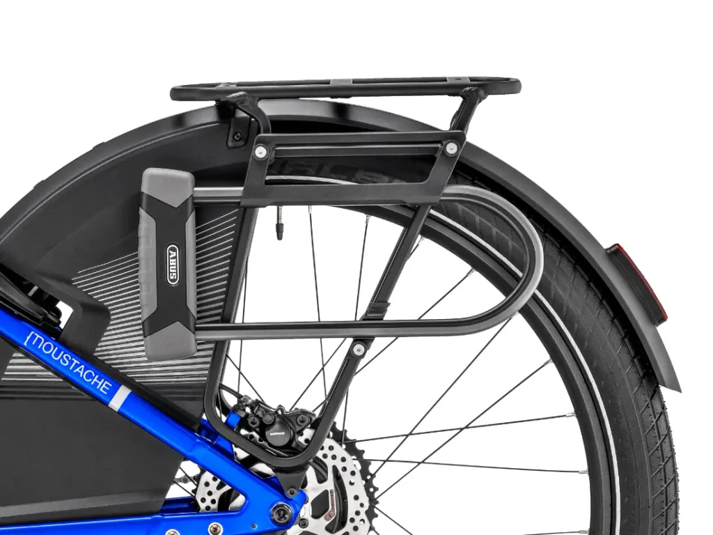 Primer plano de la rueda trasera de una bicicleta azul «Moustache Lundi 27», en el que se aprecian un portaequipajes trasero negro, un guardabarros, un freno de disco, un candado ABUS y parte del neumático: perfecta para moverse por la ciudad.