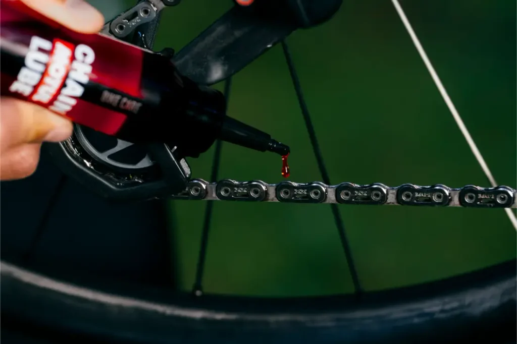 Una mano aplica lubricante para cadenas a la cadena de una bicicleta, y una gota de lubricante cae sobre los eslabones metálicos cerca de la rueda trasera.