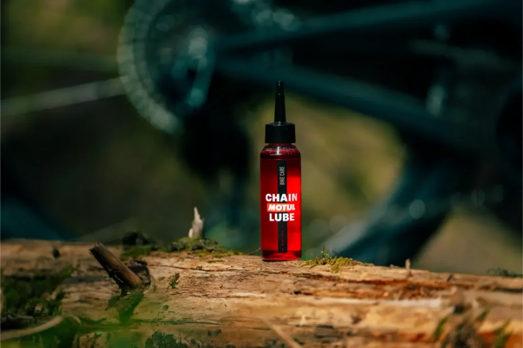 Una botella roja de lubricante para cadenas Motul está colocada en posición vertical sobre un tronco al aire libre, con una rueda de bicicleta borrosa visible al fondo.