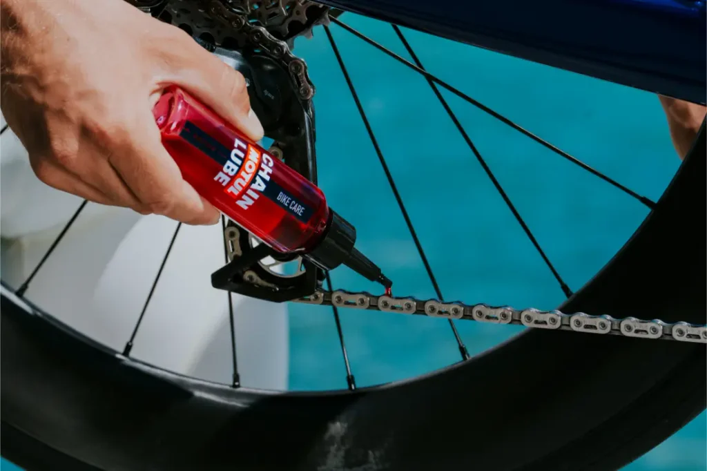 Una persona aplica lubricante rojo para cadenas, procedente de un frasco, sobre la cadena de una bicicleta; en la imagen se ven la rueda trasera y el cassette sobre un fondo azul.