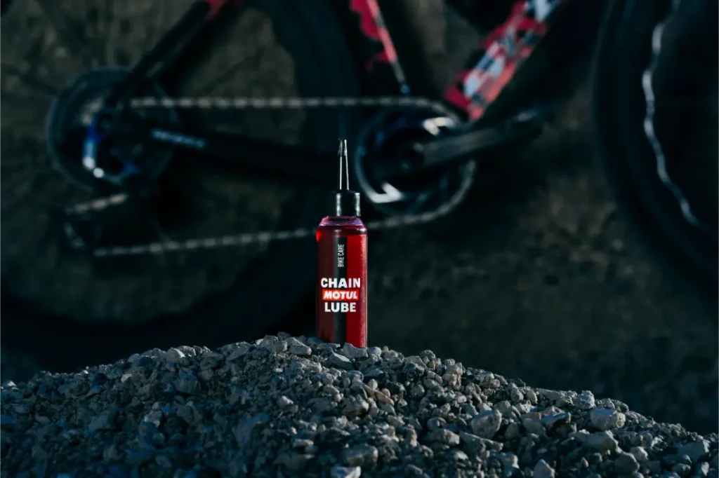 Una botella roja con la etiqueta «Chain Motus Lube» se encuentra en posición vertical sobre un montón de grava, con una rueda y un cuadro de bicicleta difuminados al fondo.